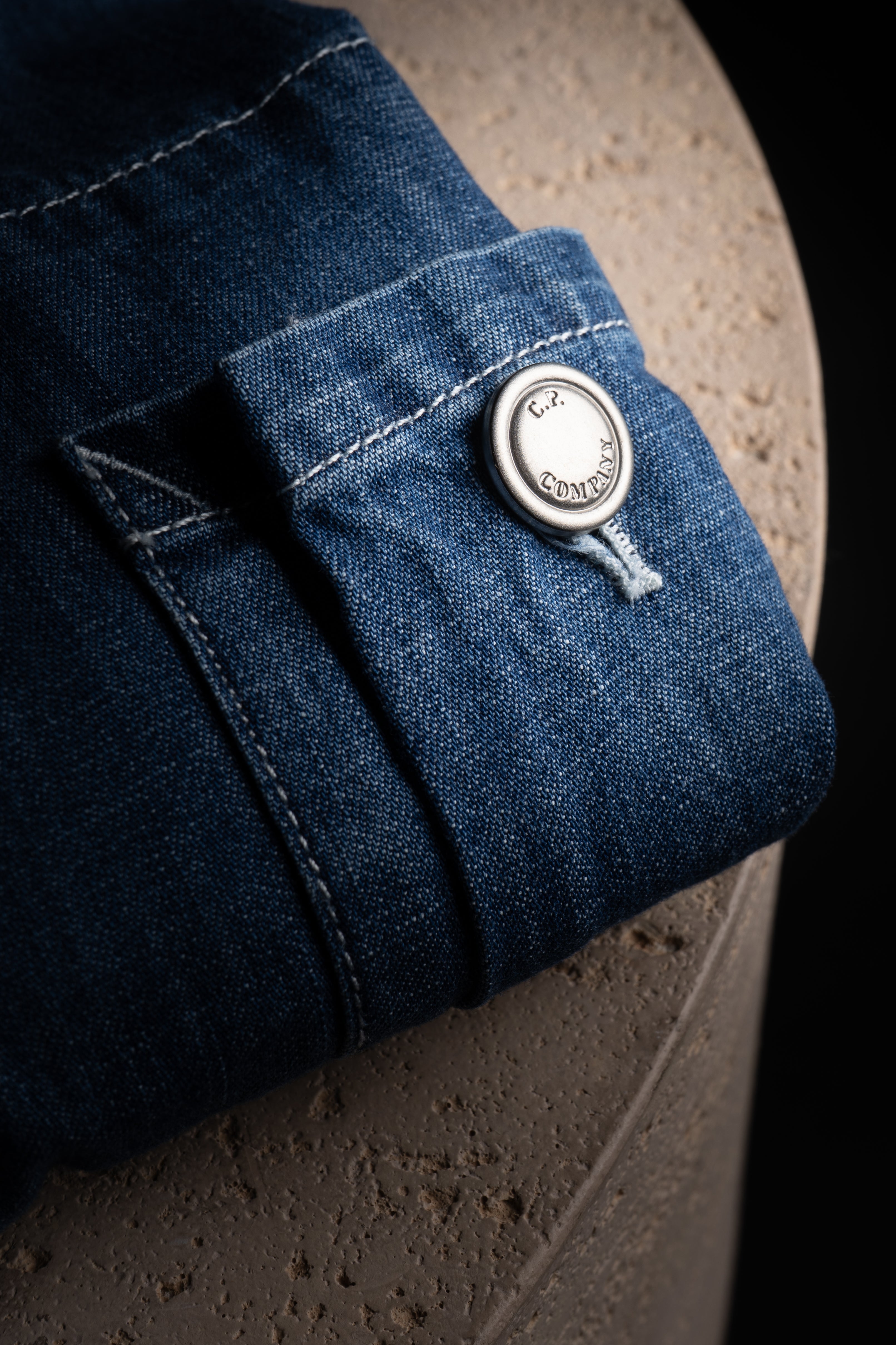 Détail de la Veste CP Company Bleu Jeans avec bouton brodé C.P. sur une surface texturée.