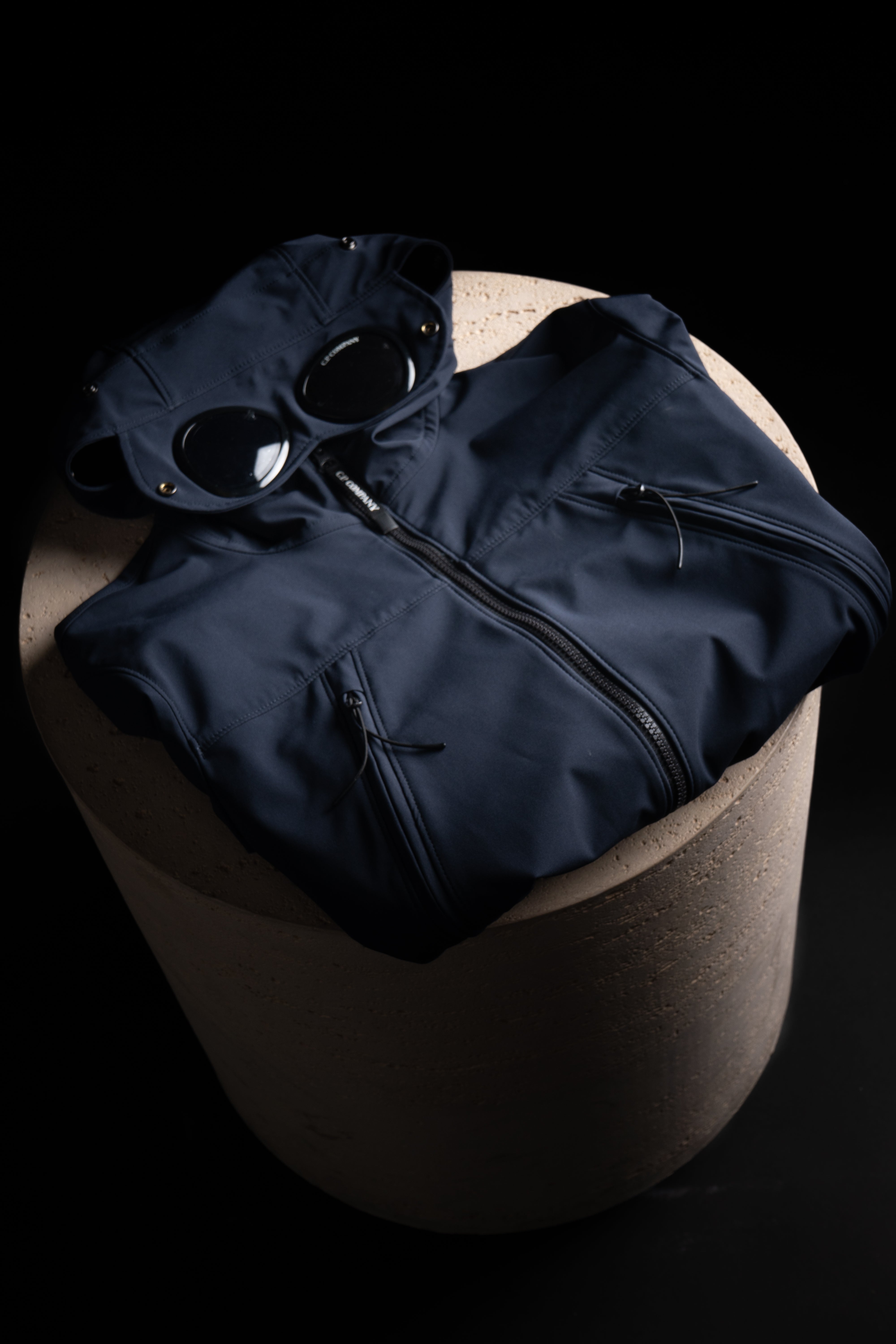 Veste CP Company bleu marine avec lunettes intégrées, sur un support en béton, offrant style et confort.
