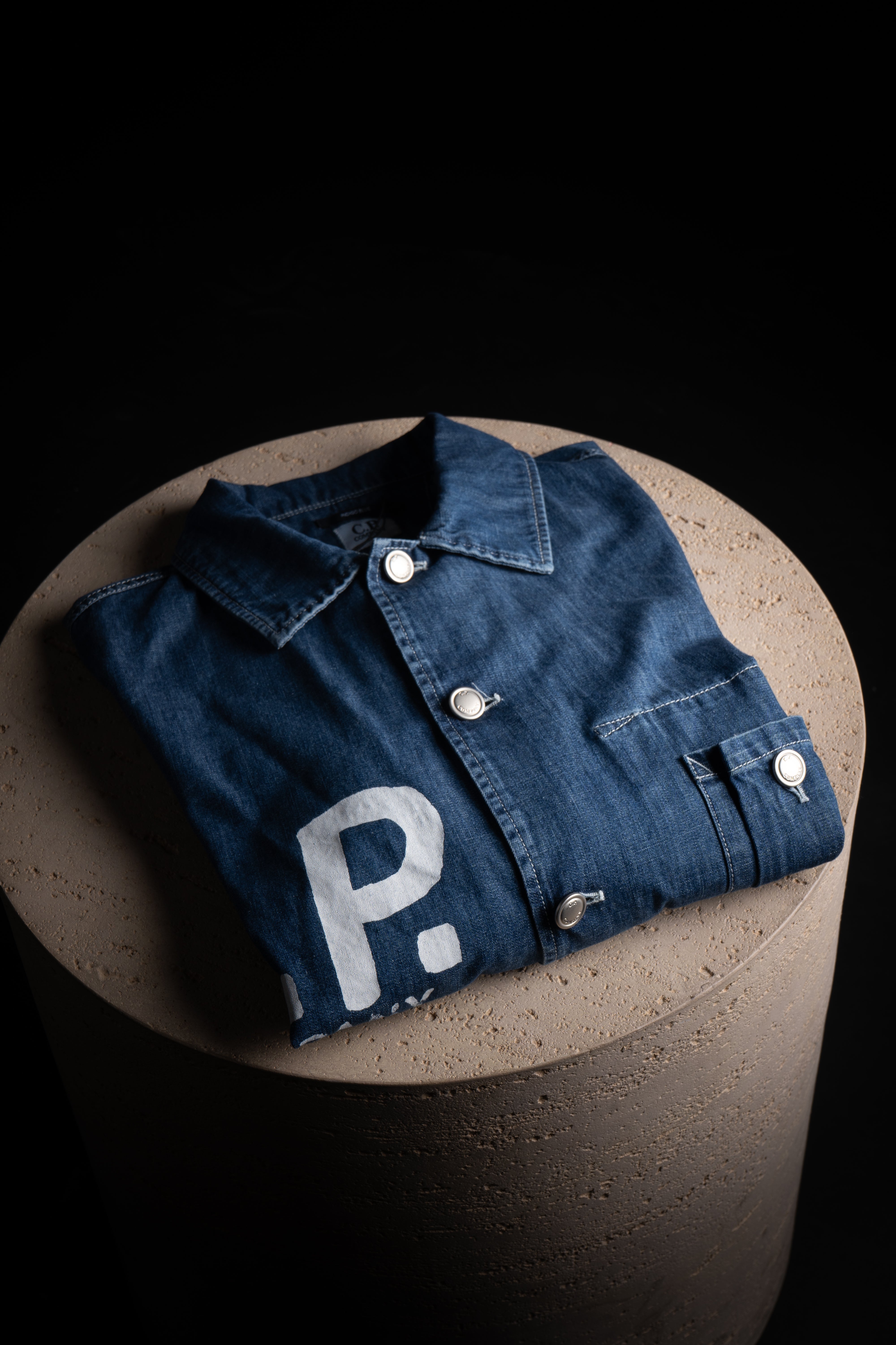 Veste CP Company Bleu Jeans en denim bleu, avec logo brodé, posée sur un support en pierre.